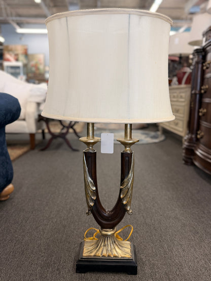 Decorative table lamp featuring a dark U-shaped body with metallic leaf accents, a fluted metallic pedestal, black square base, and a large cream-colored drum shade.