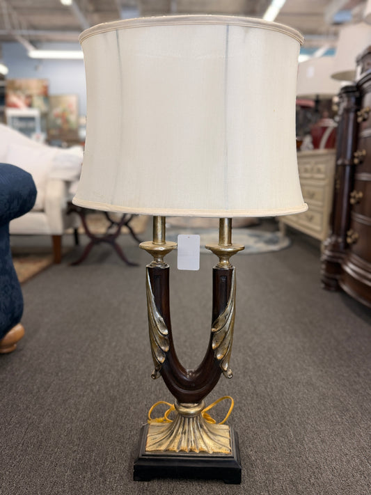 Decorative table lamp featuring a dark U-shaped body with metallic leaf accents, a fluted metallic pedestal, black square base, and a large cream-colored drum shade.