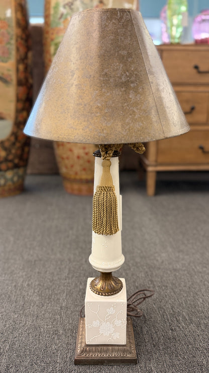 A decorative table lamp featuring a slender white floral ceramic body, a metallic-finish shade, brass accents, and a gold tassel.