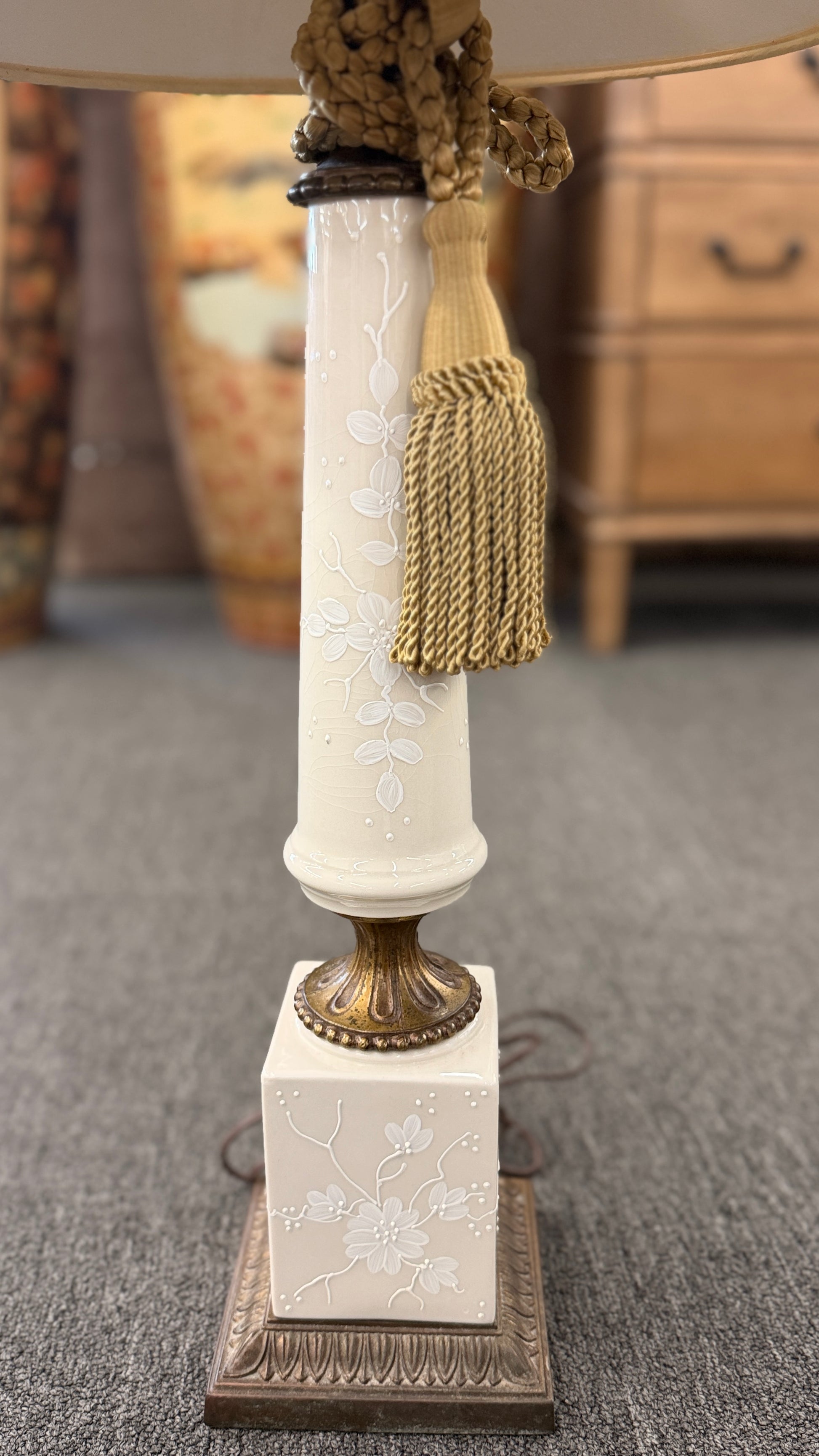 A decorative table lamp featuring a slender white floral ceramic body, a metallic-finish shade, brass accents, and a gold tassel.