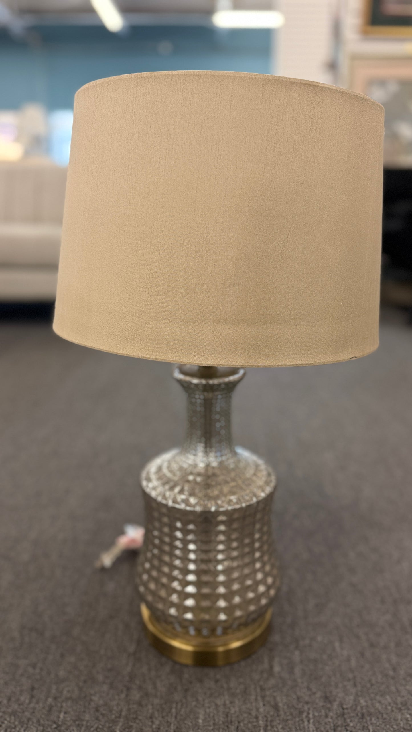 A modern transitional accent lamp featuring a diamond-quilted tinted glass urn base on a brass platform with a beige linen drum shade.
