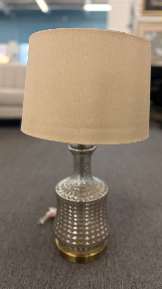 A modern transitional accent lamp featuring a diamond-quilted tinted glass urn base on a brass platform with a beige linen drum shade.