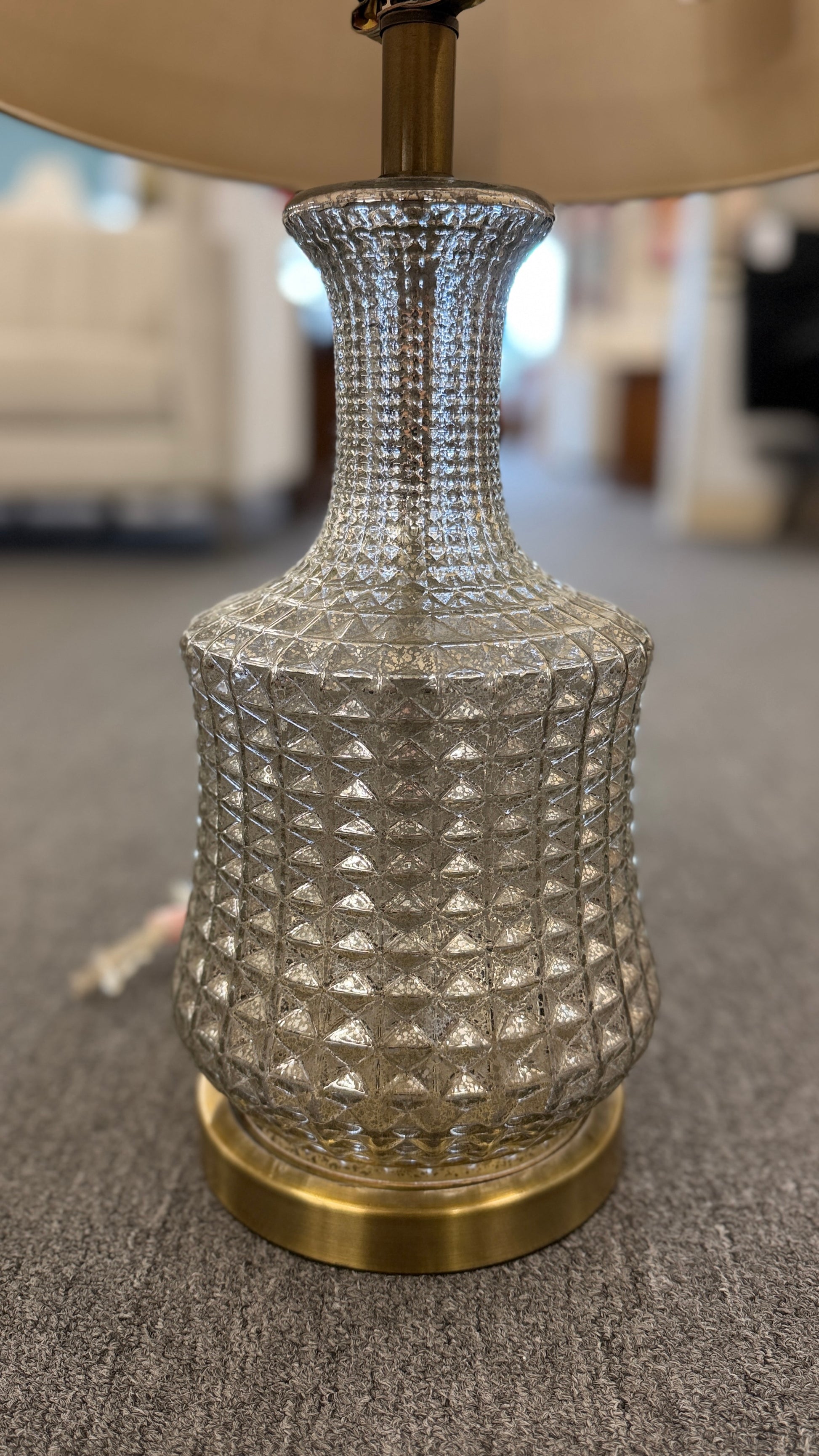 A modern transitional accent lamp featuring a diamond-quilted tinted glass urn base on a brass platform with a beige linen drum shade.