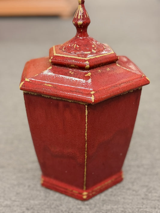 A small decorative ceramic lidded urn featuring a tapered square base, distressed red finish, gold highlights, and an ornate finial.