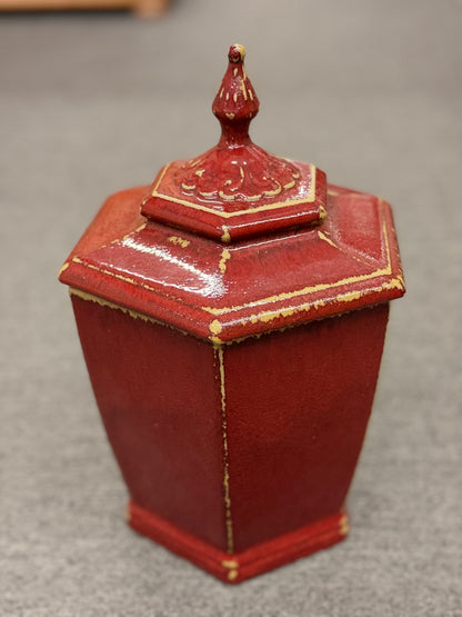 A small decorative ceramic lidded urn featuring a tapered square base, distressed red finish, gold highlights, and an ornate finial.