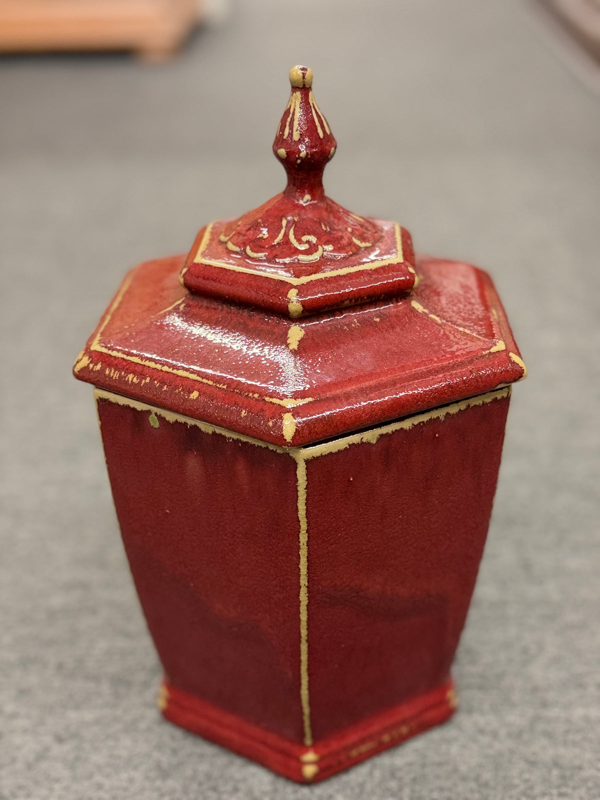 A small decorative ceramic lidded urn featuring a tapered square base, distressed red finish, gold highlights, and an ornate finial.