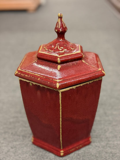 A small decorative ceramic lidded urn featuring a tapered square base, distressed red finish, gold highlights, and an ornate finial.