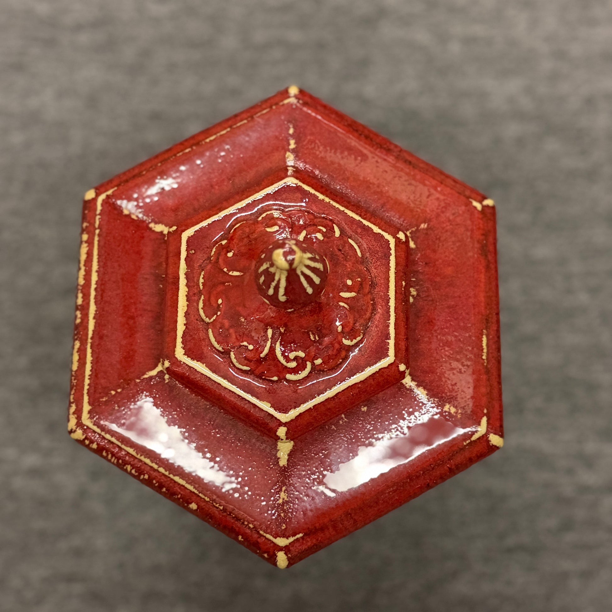 A small decorative ceramic lidded urn featuring a tapered square base, distressed red finish, gold highlights, and an ornate finial.