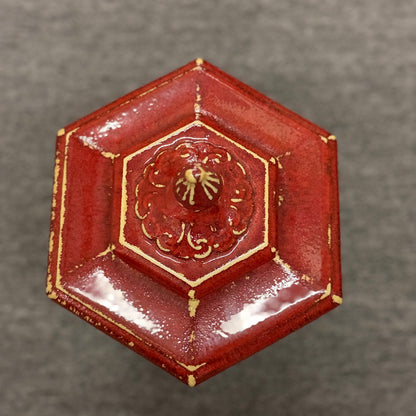 A small decorative ceramic lidded urn featuring a tapered square base, distressed red finish, gold highlights, and an ornate finial.