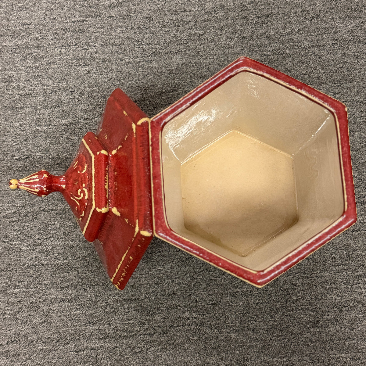 A small decorative ceramic lidded urn featuring a tapered square base, distressed red finish, gold highlights, and an ornate finial.
