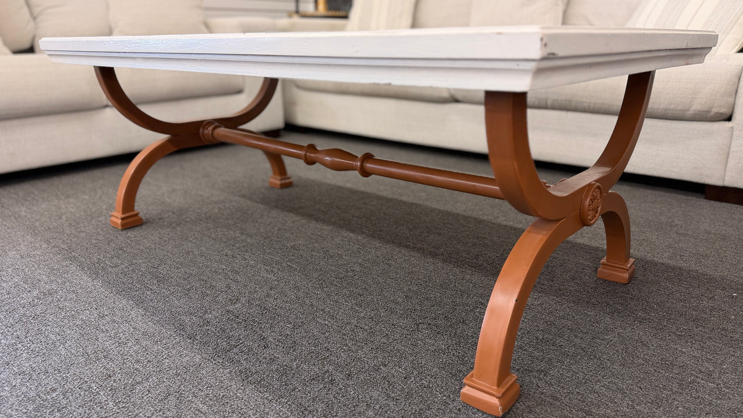 A rectangular coffee table featuring a distressed painted white top supported by an arched reddish-brown wood base with a central stretcher.