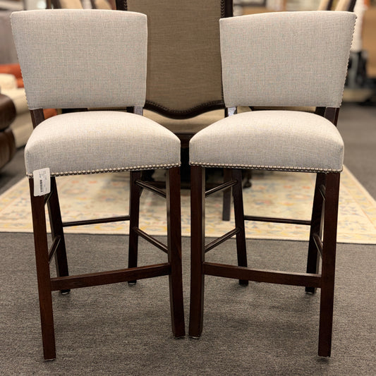A pair of upholstered high-back counter stools featuring light grey textured fabric, classic metal nailhead trim, and dark-stained wooden legs with footrests.