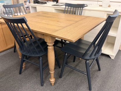 A farmhouse-style dining set featuring a rectangular light-wood extension table and four dark painted wooden Windsor-style chairs.