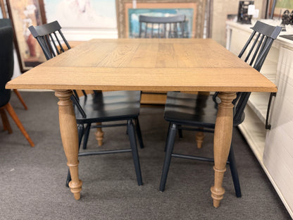 A farmhouse-style dining set featuring a rectangular light-wood extension table and four dark painted wooden Windsor-style chairs.