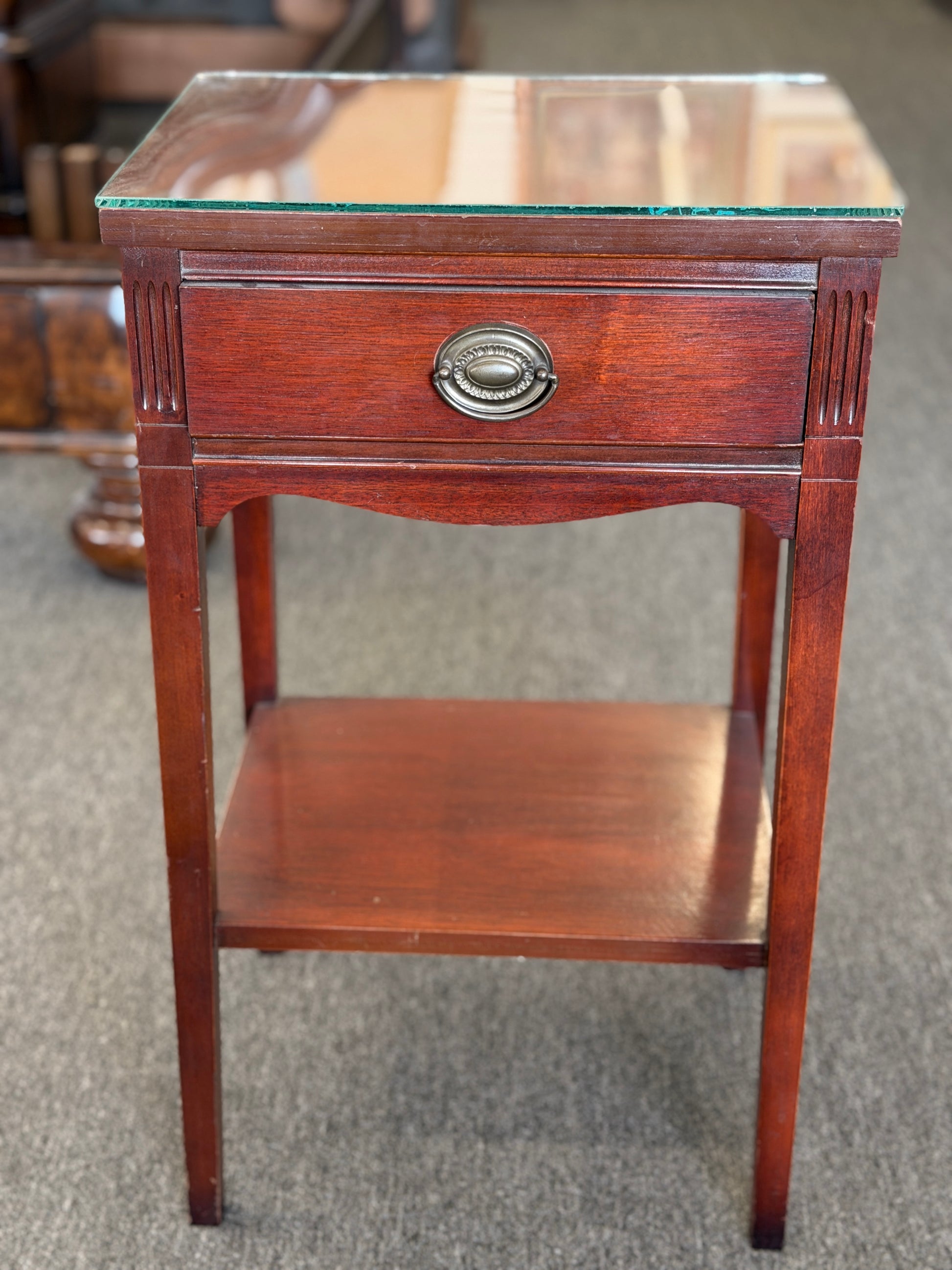 A Federal-style wood accent table featuring fluted corner accents, an arched apron, an oval rosette drop pull, an open lower shelf, and a glass top in good condition.