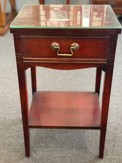 A compact Federal-style wood end table featuring fluted corner carvings, a single drawer with brass bail pull, lower shelf, and a custom glass top in good condition.