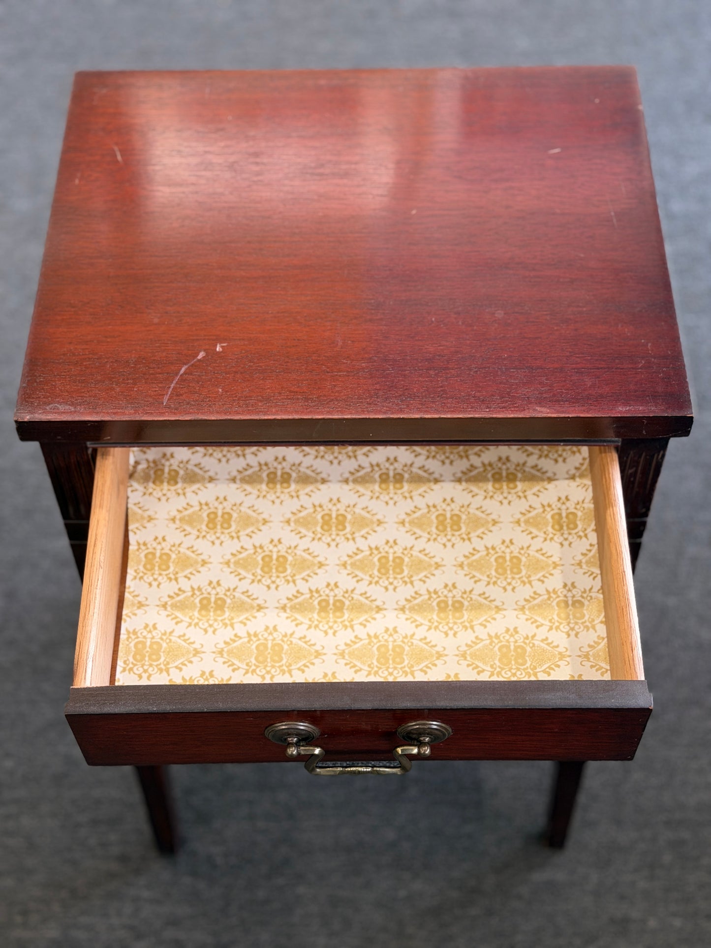 A compact Federal-style wood end table featuring fluted corner carvings, a single drawer with brass bail pull, lower shelf, and a custom glass top in good condition.