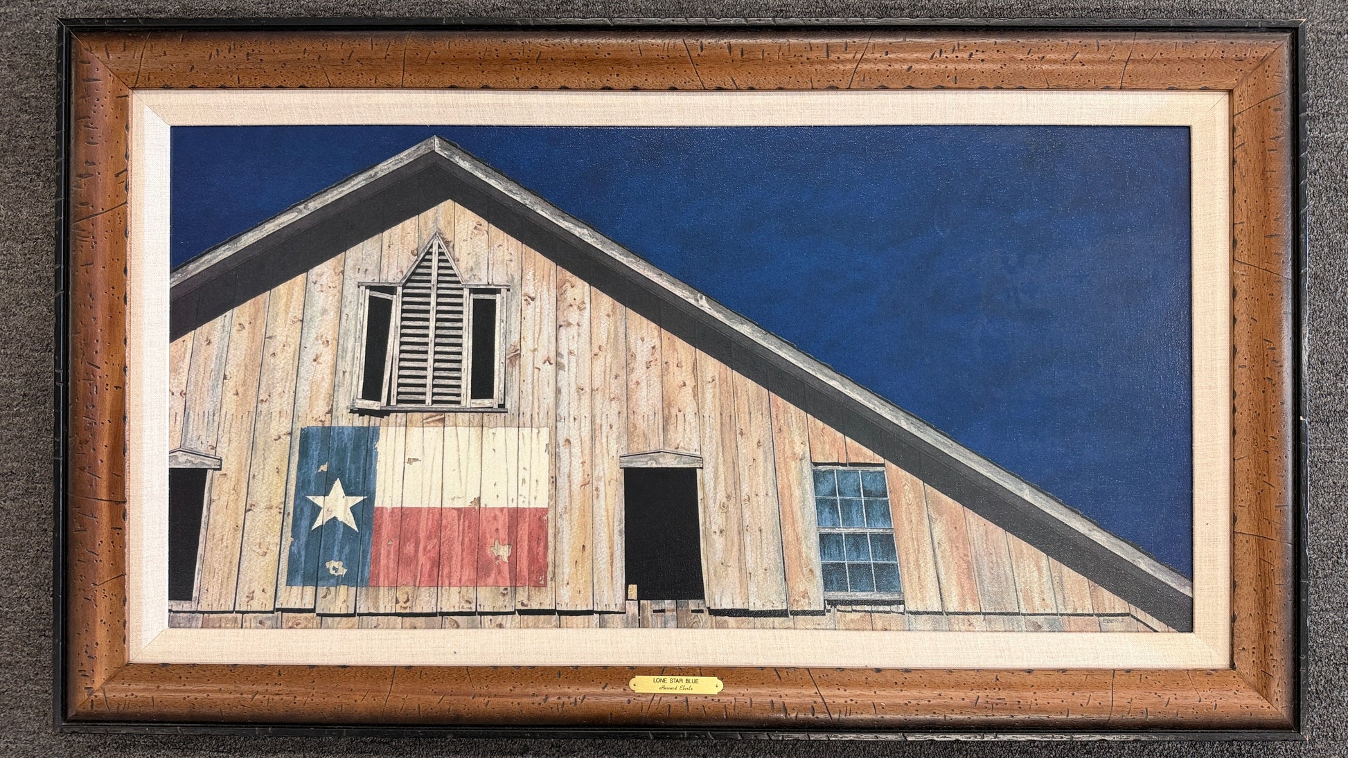 A wide framed rustic art print depicting a weathered wooden barn painted with the Texas state flag set in a textured wood frame with linen liner in excellent condition.