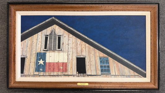 A wide framed rustic art print depicting a weathered wooden barn painted with the Texas state flag set in a textured wood frame with linen liner in excellent condition.