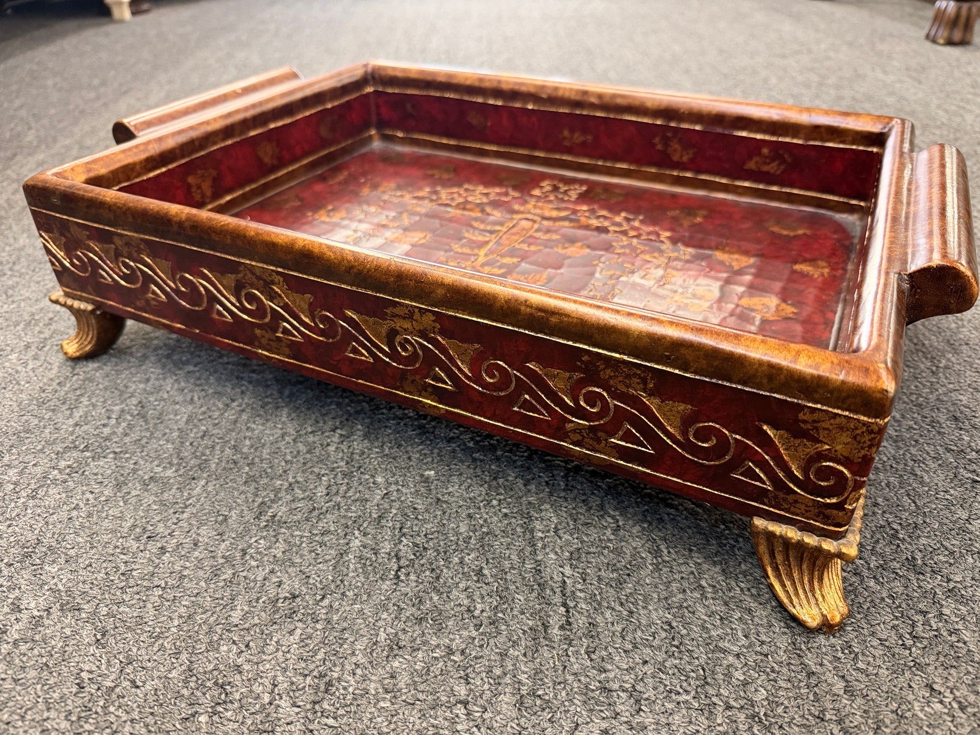 A rectangular wooden decorative vanity tray with ornate gold wave patterns on the sides, handles, short feet, and a faux-tortoiseshell interior with gold botanical designs.
