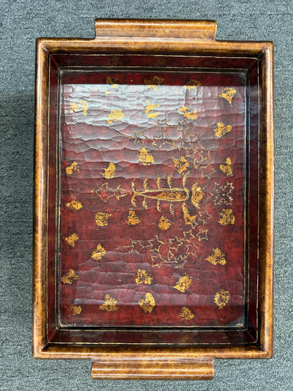 A rectangular wooden decorative vanity tray with ornate gold wave patterns on the sides, handles, short feet, and a faux-tortoiseshell interior with gold botanical designs.