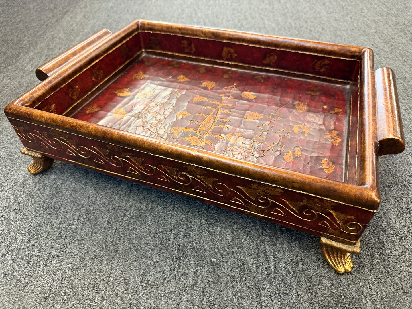 A rectangular wooden decorative vanity tray with ornate gold wave patterns on the sides, handles, short feet, and a faux-tortoiseshell interior with gold botanical designs.