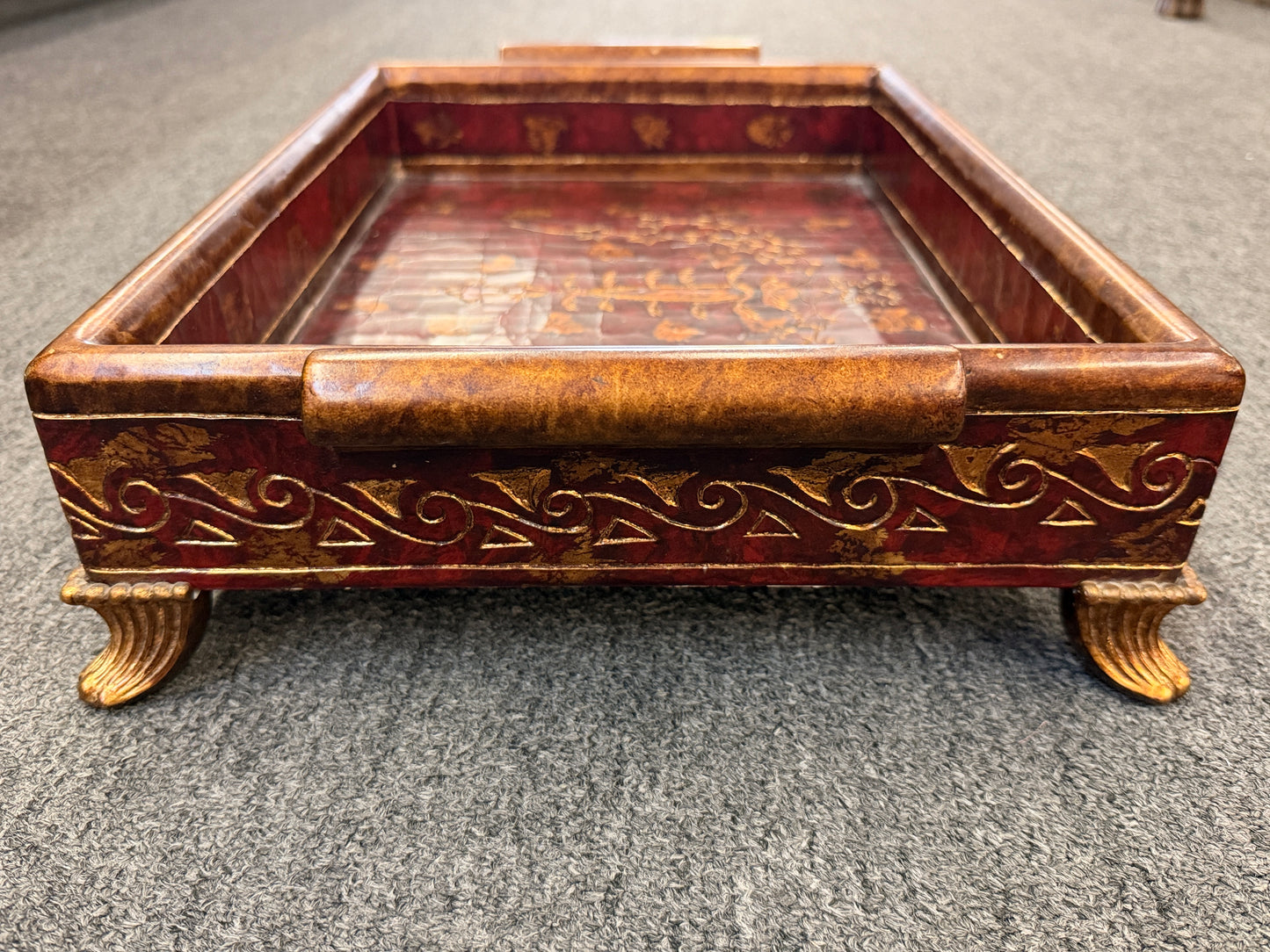 A rectangular wooden decorative vanity tray with ornate gold wave patterns on the sides, handles, short feet, and a faux-tortoiseshell interior with gold botanical designs.