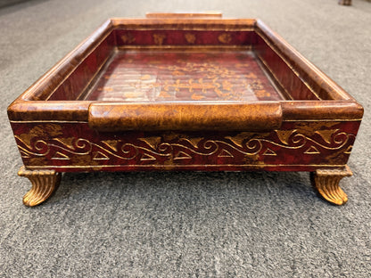 A rectangular wooden decorative vanity tray with ornate gold wave patterns on the sides, handles, short feet, and a faux-tortoiseshell interior with gold botanical designs.