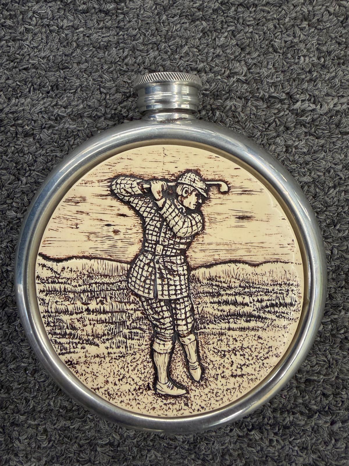 A circular pocket hip flask featuring a central scrimshaw-style engraving of a golfer mid-swing in excellent condition.