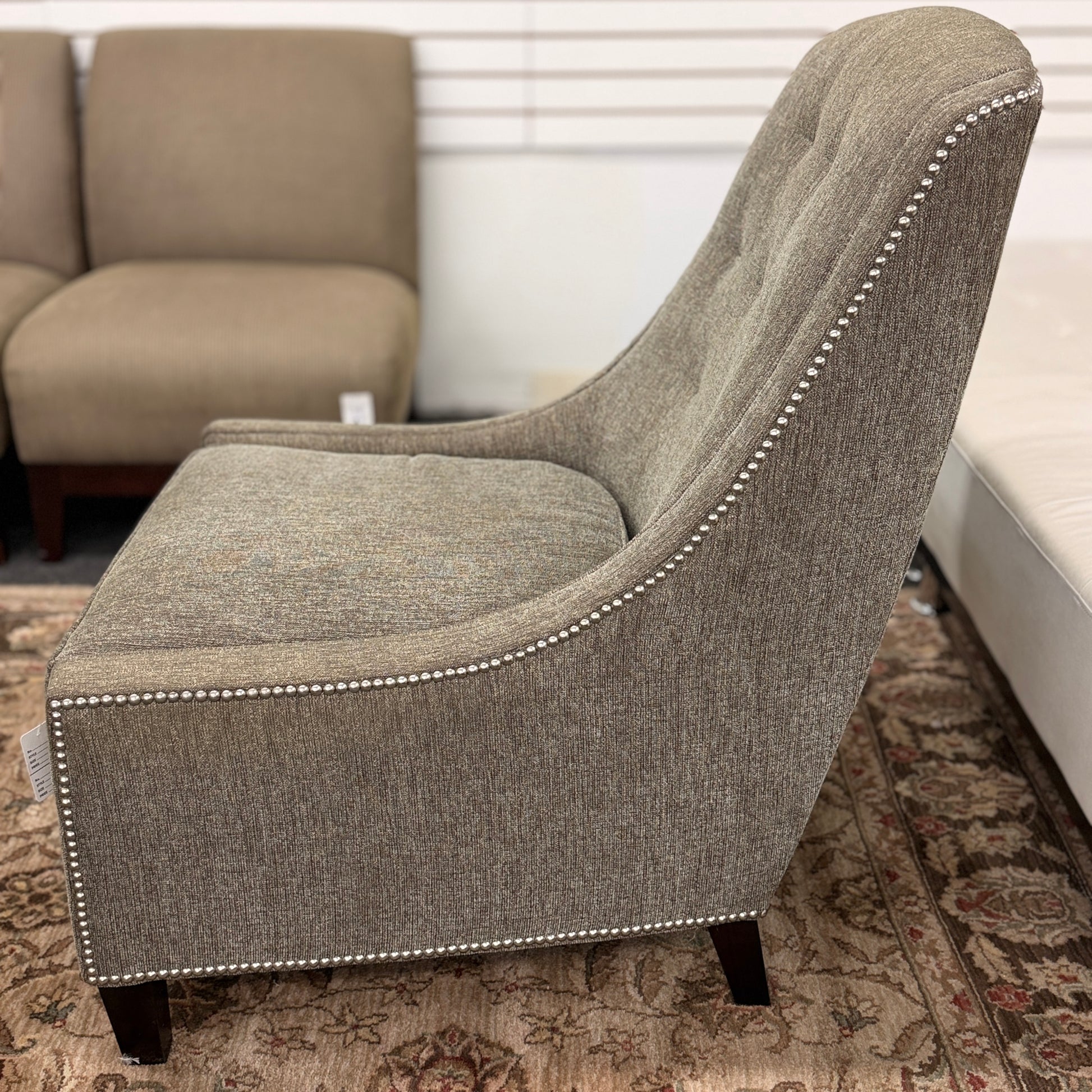 Gray upholstered armless accent chair featuring textured fabric, a gently curved tufted back, silver nailhead trim, and dark wooden legs.