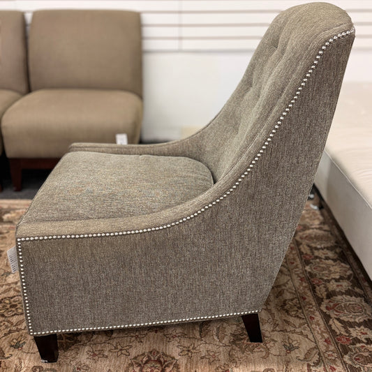 Gray upholstered armless accent chair featuring textured fabric, a gently curved tufted back, silver nailhead trim, and dark wooden legs.