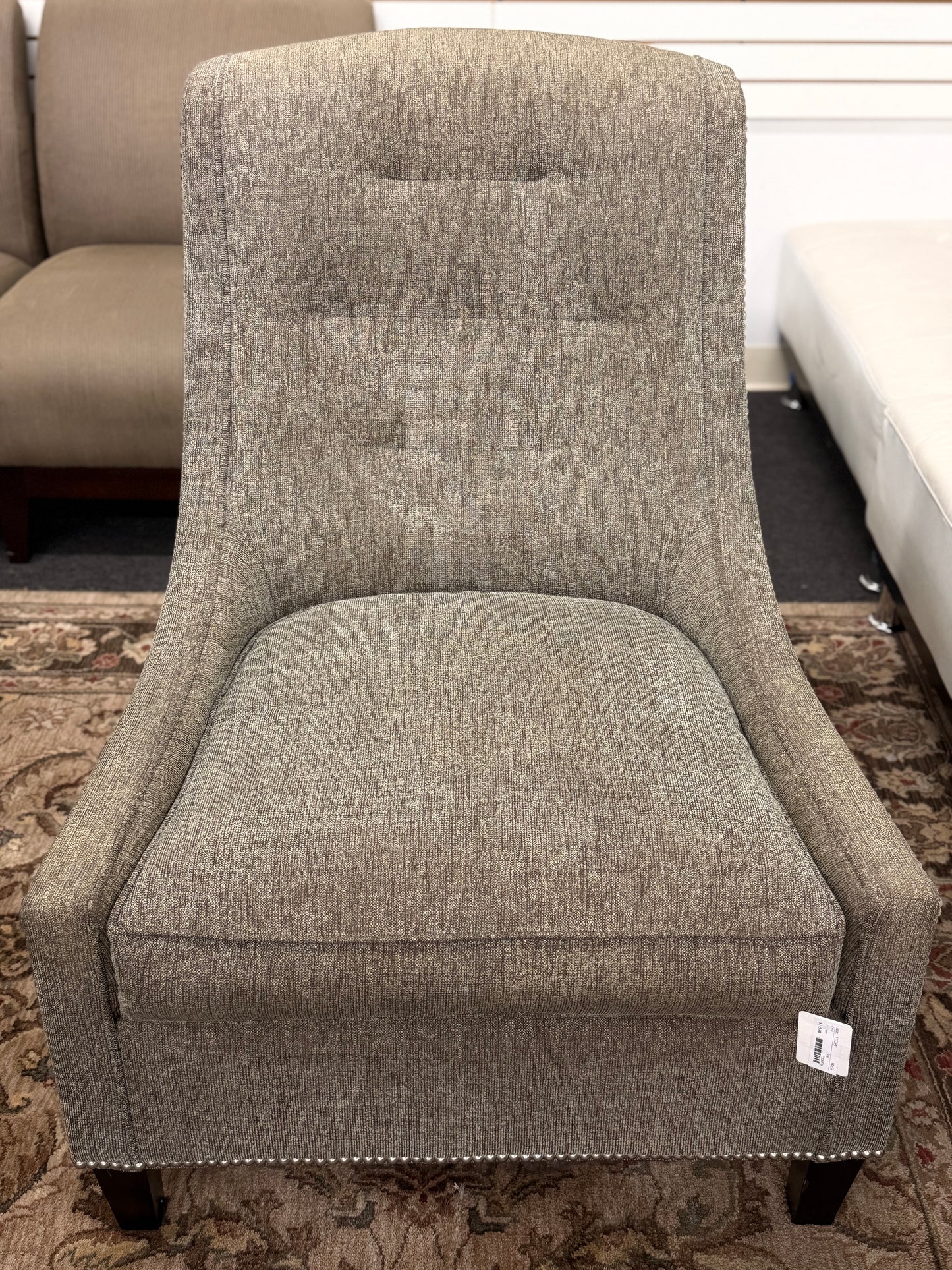 Gray upholstered armless accent chair featuring textured fabric, a gently curved tufted back, silver nailhead trim, and dark wooden legs.