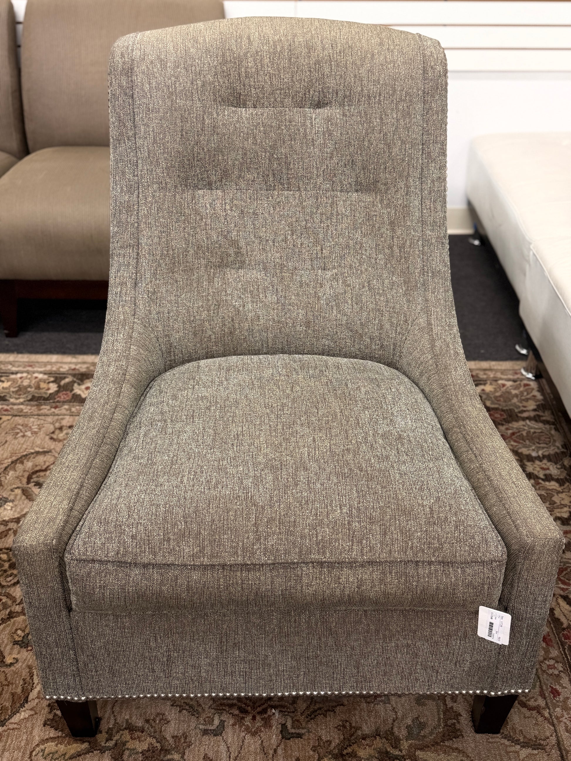 Gray upholstered armless accent chair featuring textured fabric, a gently curved tufted back, silver nailhead trim, and dark wooden legs.