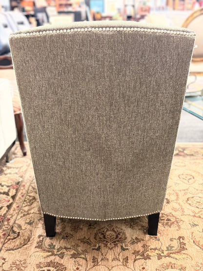 Gray upholstered armless accent chair featuring textured fabric, a gently curved tufted back, silver nailhead trim, and dark wooden legs.
