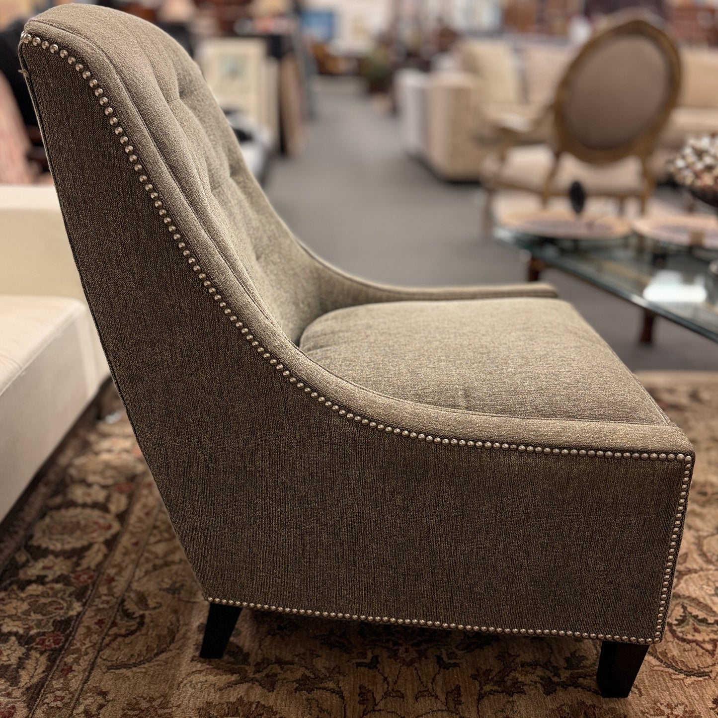 Gray upholstered armless accent chair featuring textured fabric, a gently curved tufted back, silver nailhead trim, and dark wooden legs.