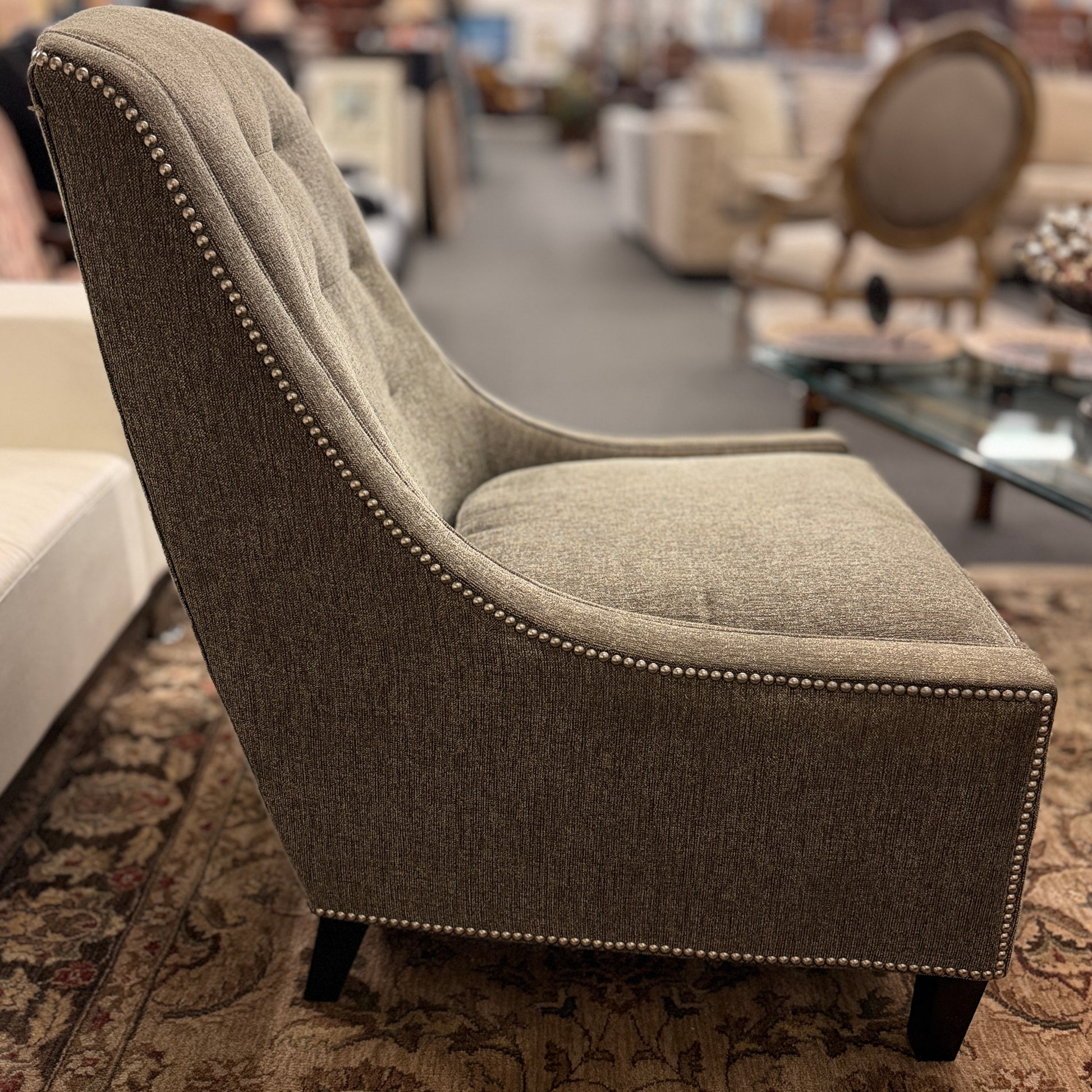 Gray upholstered armless accent chair featuring textured fabric, a gently curved tufted back, silver nailhead trim, and dark wooden legs.