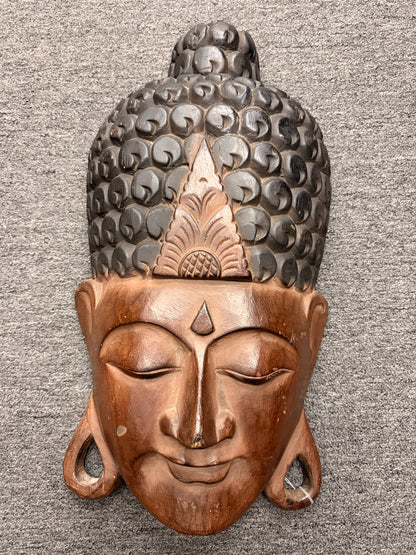 A hand-carved wooden Buddha wall mask finished in two-tone brown and black tones with an ornate headdress in excellent condition.