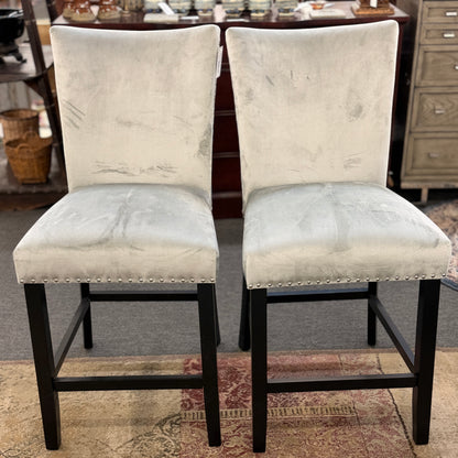 A pair of counter-height bar stools upholstered in light gray velvet-like fabric with silver nailhead trim and dark square wooden legs.