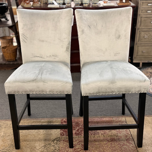 A pair of counter-height bar stools upholstered in light gray velvet-like fabric with silver nailhead trim and dark square wooden legs.