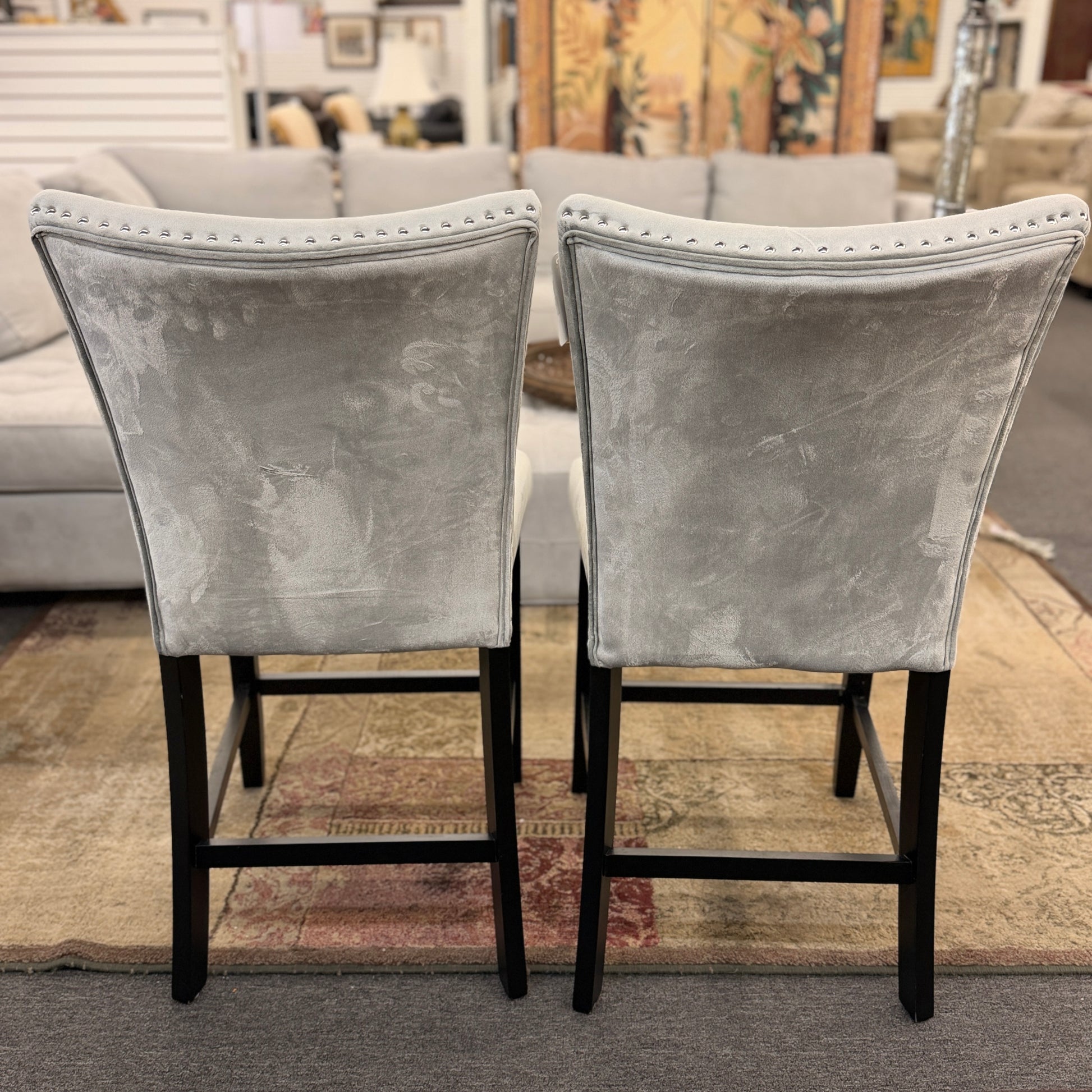 A pair of counter-height bar stools upholstered in light gray velvet-like fabric with silver nailhead trim and dark square wooden legs.