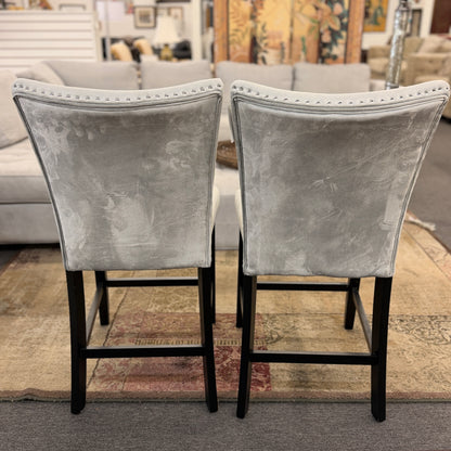 A pair of counter-height bar stools upholstered in light gray velvet-like fabric with silver nailhead trim and dark square wooden legs.