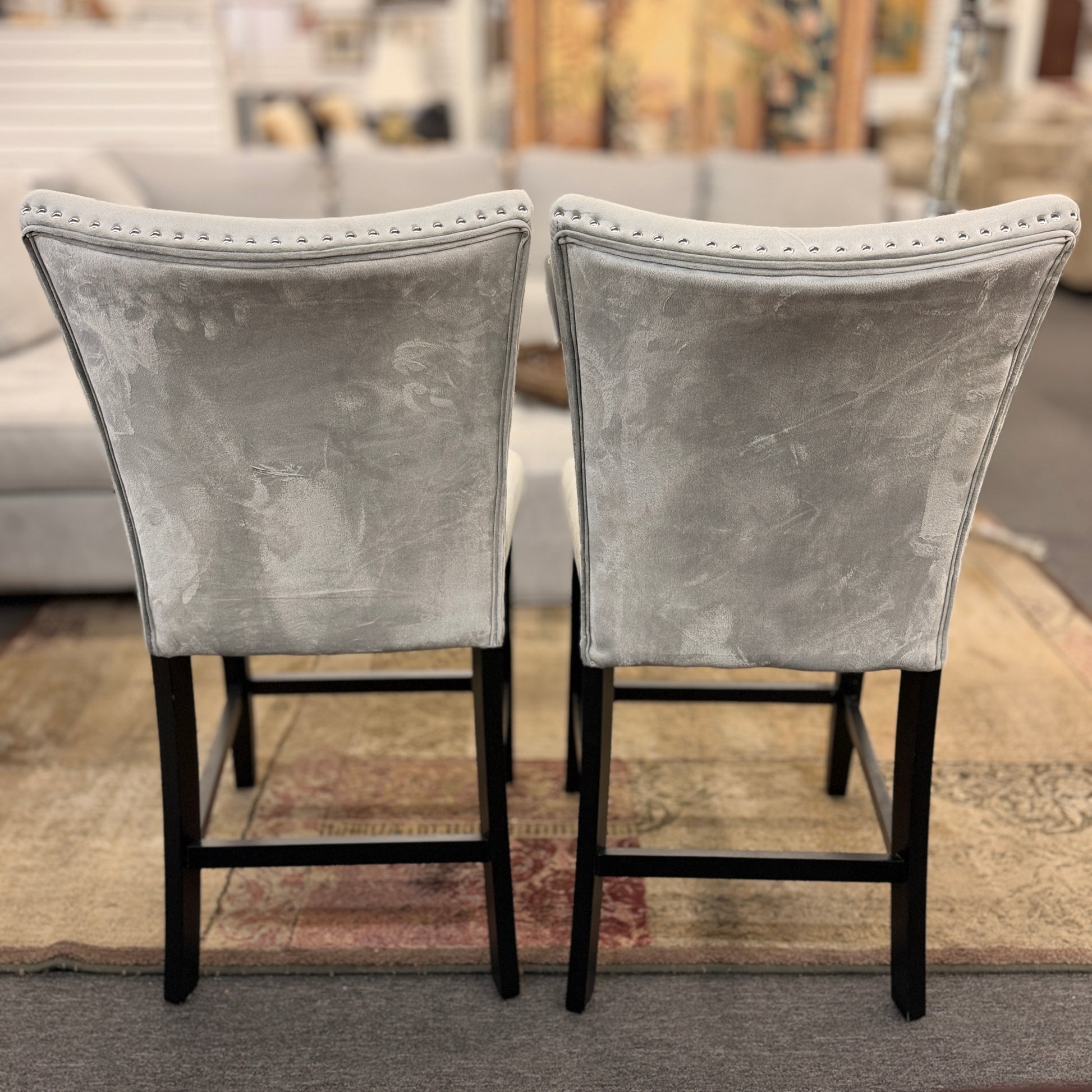 A pair of counter-height bar stools upholstered in light gray velvet-like fabric with silver nailhead trim and dark square wooden legs.