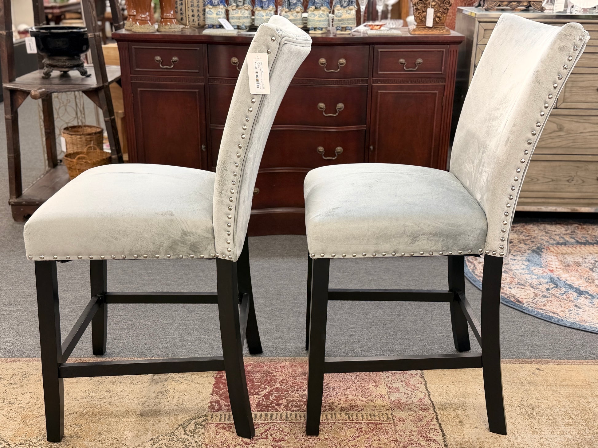 A pair of counter-height bar stools upholstered in light gray velvet-like fabric with silver nailhead trim and dark square wooden legs.