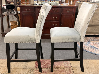 A pair of counter-height bar stools upholstered in light gray velvet-like fabric with silver nailhead trim and dark square wooden legs.