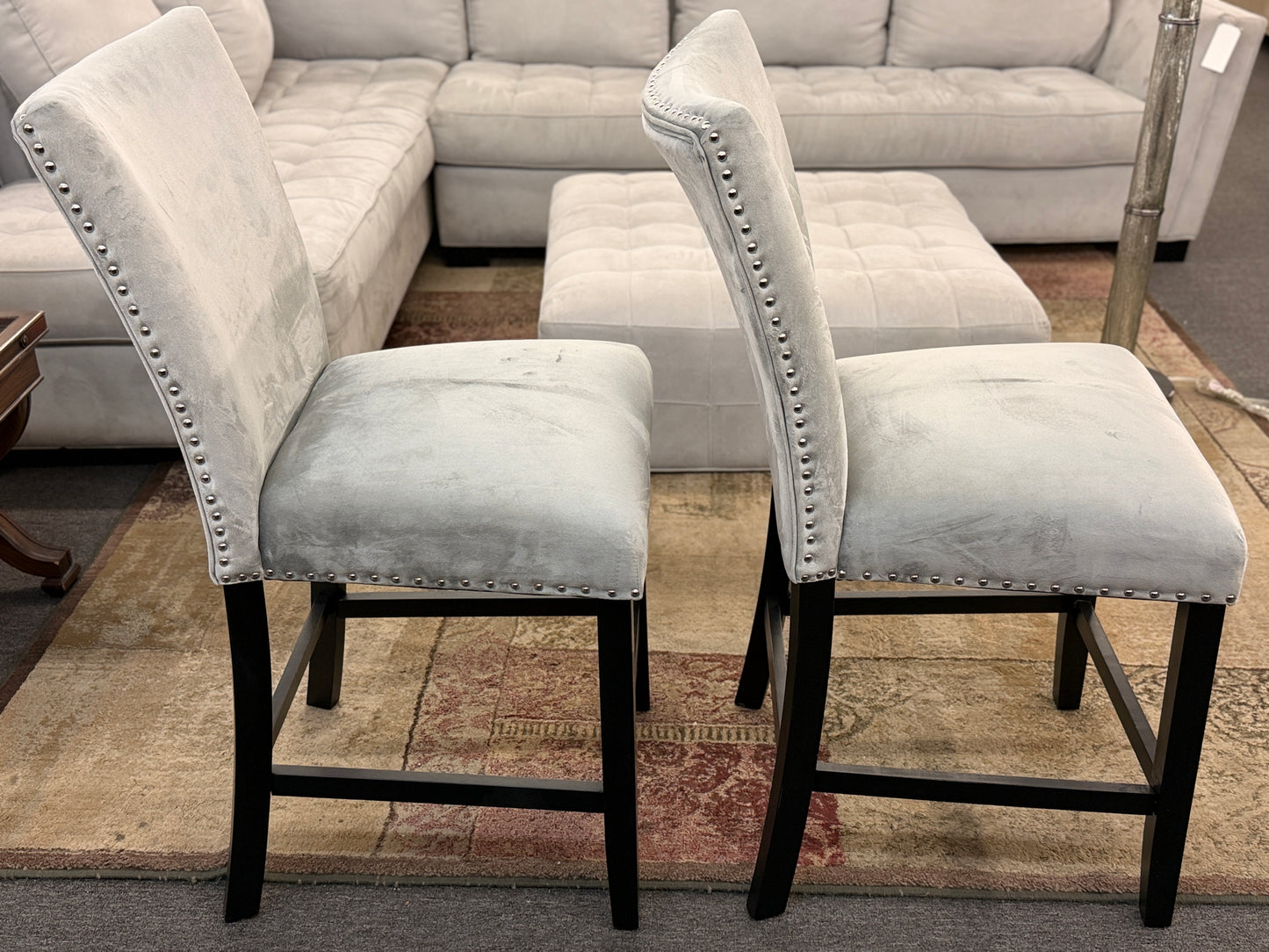 A pair of counter-height bar stools upholstered in light gray velvet-like fabric with silver nailhead trim and dark square wooden legs.