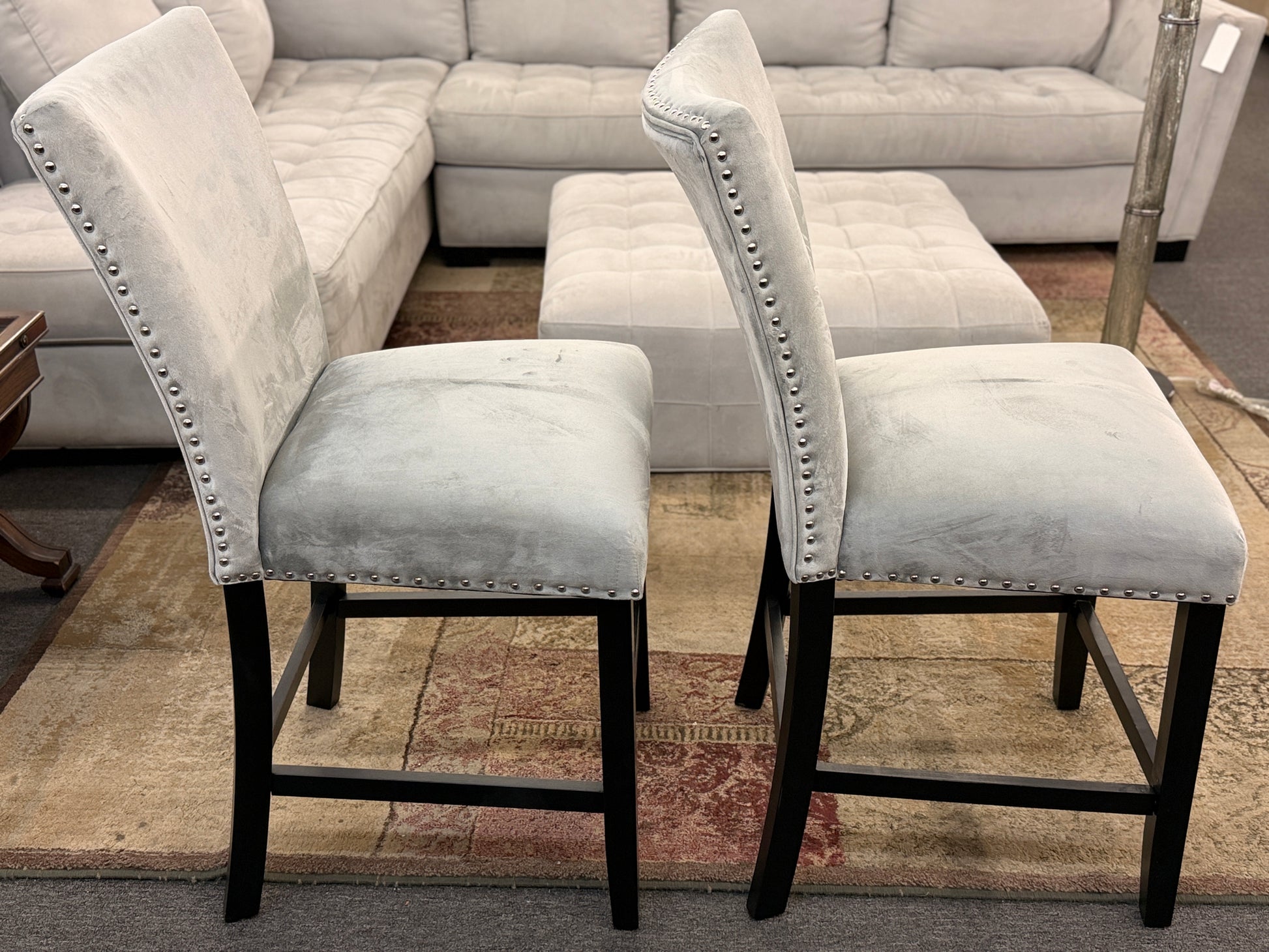 A pair of counter-height bar stools upholstered in light gray velvet-like fabric with silver nailhead trim and dark square wooden legs.