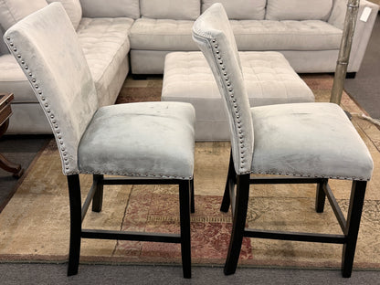 A pair of counter-height bar stools upholstered in light gray velvet-like fabric with silver nailhead trim and dark square wooden legs.