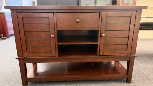 A medium-brown wooden console server featuring a central drawer, open center cubby, two louvered side doors, and a full-length lower display shelf.