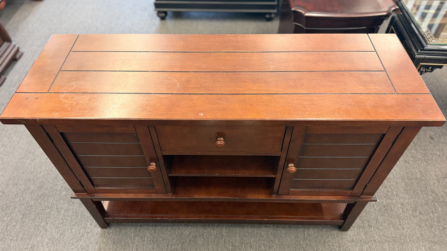 A medium-brown wooden console server featuring a central drawer, open center cubby, two louvered side doors, and a full-length lower display shelf.