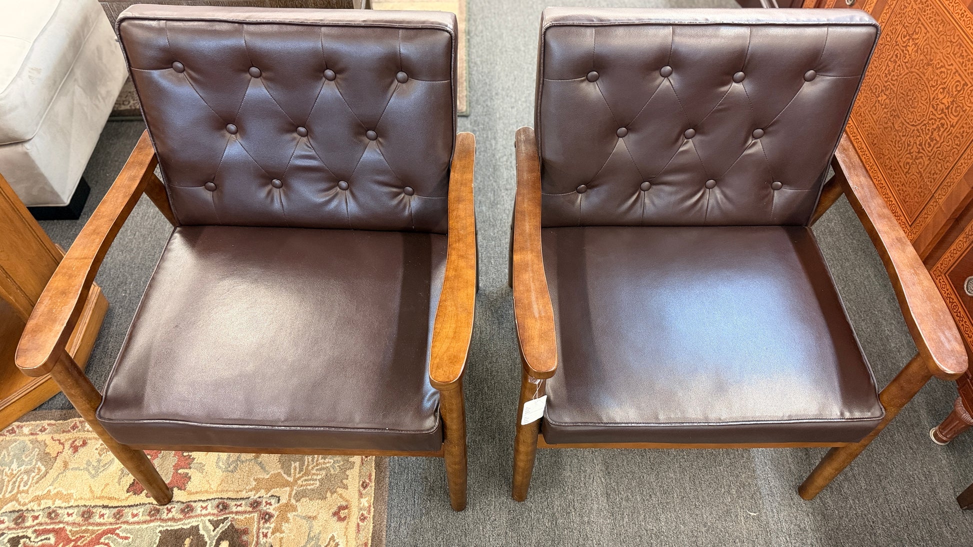 A pair of mid-century modern style accent armchairs featuring dark brown button-tufted backrests, smooth seat cushions, and exposed medium-toned wooden frames with tapered legs.